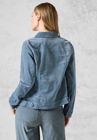 Veste en chambray bleu avec un col, des détails de boutons aux poignets et à l'ourlet, et une coupe décontractée. Texture lisse et tissu léger.
