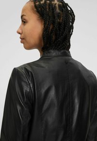Giacca di pelle nera con una superficie liscia, design su misura e un piccolo bottone sul retro. Le cuciture a vista completano la struttura.