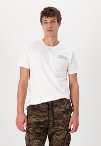 Hvid bomulds t-shirt med korte ærmer og et grønt grafisk print på brystet. Kombineret med brune cargo bukser i camouflage mønster med lommer.