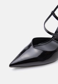 Chaussure pointue en cuir verni noir avec fine bride à la cheville et boucle, présentée sur fond blanc.