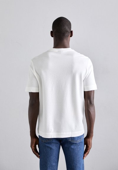 T-shirt blanc à manches courtes avec un motif texturé, col rond et fentes sur les côtés, associé à un jean en denim bleu.