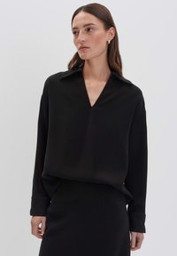 Donna con lunghi capelli castani, che indossa una blusa nera a V con colletto e maniche lunghe, abbinata a una gonna nera, su uno sfondo chiaro e semplice.