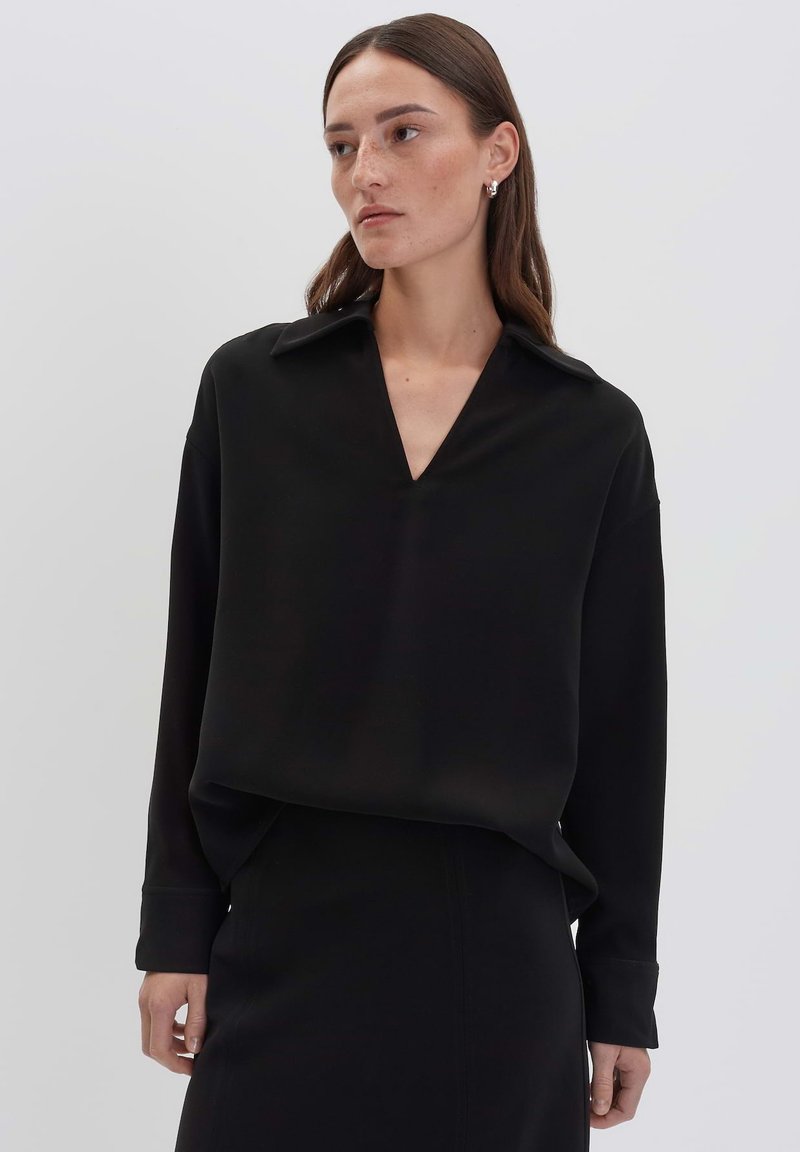 Donna con lunghi capelli castani, che indossa una blusa nera a V con colletto e maniche lunghe, abbinata a una gonna nera, su uno sfondo chiaro e semplice.