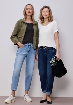 Zwei Frauen stehen vor einer schlichten Wand, eine trägt eine grüne Jacke, ein schwarzes Top, helle Jeans und weiße Sneaker, die andere ein weißes T-Shirt, dunkle Jeans und schwarze Ballerinas.