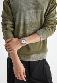 Olive green sweater with a textured finish and ribbed cuffs, paired with a clear wristwatch featuring a simple white face and black markings.