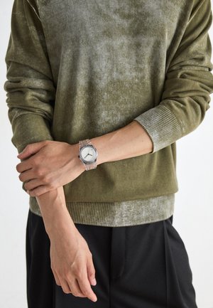 Pull en vert olive avec une finition texturée et des poignets côtelés, associé à une montre-bracelet transparente avec un cadran blanc simple et des marquages noirs.