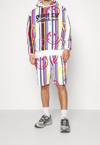 Kleurrijke hoodie en shortset, met verticale strepen in rood, geel, blauw en wit, met glimlachende gezichten en logodetails.