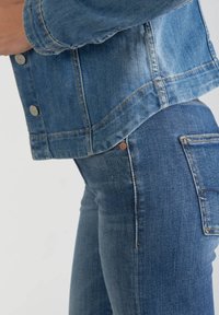 La giacca di jeans presenta un lavaggio azzurro chiaro con cuciture arancioni a contrasto e bottoni a pressione argentati, abbinata a jeans attillati blu scuro.
