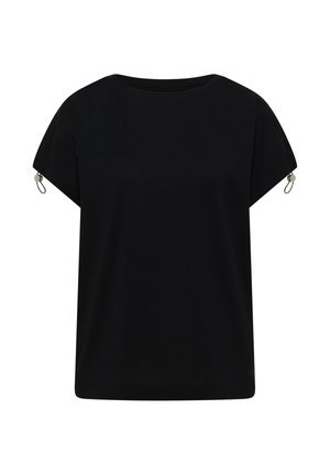 Femme souriante marchant à l'intérieur, portant un t-shirt noir à manches courtes, un short en jean bleu clair et des chaussures beiges sans lacets, sur un fond neutre.