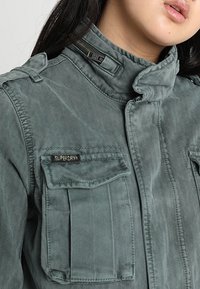 Grijze katoenen jas met een hoge kraag, ritssluiting en twee voorzakken met klep. Bevat een klein "SUPERDRY" logo op de linkse zak.