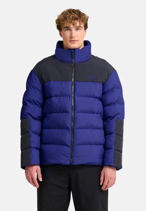 Junger Mann mit einer blau-schwarzen gesteppte Winterjacke mit hohem Kragen, stehend vor einem einfarbigen hellen Hintergrund.