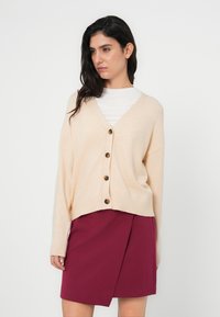 Ανοιχτό μπεζ ζακέτα με V-λαιμόκοψη, μακριά μανίκια και ξύλινα κουμπιά, σε συνδυασμό με φούστα τύπου wrap σε μπορντό. Λείο ύφασμα, άνετη εφαρμογή.