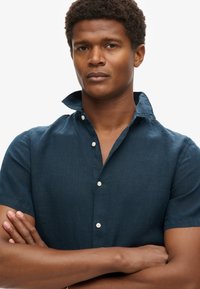 Camicia in lino blu scuro a maniche corte con colletto button-down, caratterizzata da una finitura testurizzata e bottoni bianchi. Il modello ha le braccia incrociate.