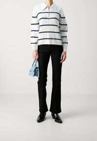 Weiß-blau gestreifter Strickpullover mit einem Reißverschlusskragen, kombiniert mit schwarzen, weit ausgestellten Hosen. Hält eine kleine hellblaue Handtasche. Schwarze Ankle-Boots.