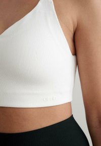 Biely rebrovaný crop top s jedným ramienkom, s jemným logom na leme a hladkou textúrou. Skombinovaný s tmavozelenými rebrovanými nohavicami.