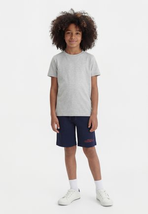 Dziecko o kręconych włosach, ubrane w jasnoszarą koszulkę, granatowe szorty z czerwonym napisem, białe skarpetki i białe trampki, stojące na białym tle.