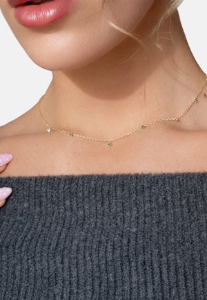 Close-up of a woman’s neck wearing a delicate gold chain necklace with small green gemstones, paired with a dark gray knitted sweater.
