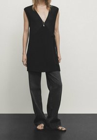 Persoon in een mouwloze zwarte V-hals tuniek, donkere wijde jeans, zwarte open teen sandalen en een hangerketting, staande tegen een effen achtergrond.