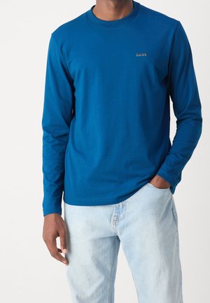 Homme portant une chemise bleue à manches longues avec le logo "BOSS" sur la poitrine et un jean bleu clair, une main dans la poche, sur un fond uni.