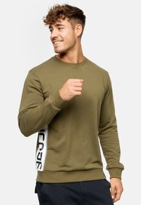 Sweatshirt vert olive avec des manches longues, un col rond et une bande latérale blanche avec du texte noir. Fabriqué à partir d'un tissu doux et texturé.