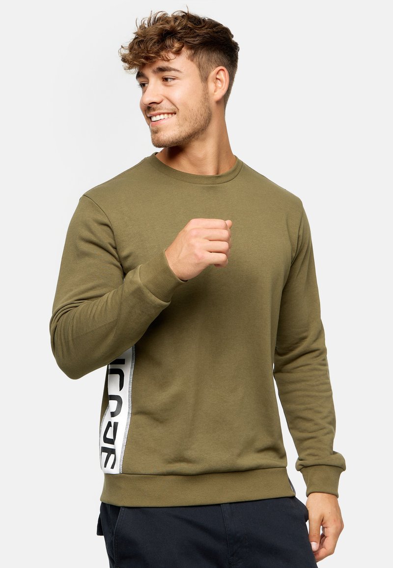 Sweatshirt vert olive avec des manches longues, un col rond et une bande latérale blanche avec du texte noir. Fabriqué à partir d'un tissu doux et texturé.