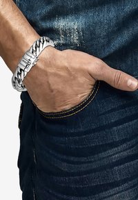 Silvermetallarmband med flätad design, utrustad med ett spänne. Bärs på en hand, med mörka denimbyxor synliga i bakgrunden.