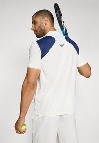 Λευκό αθλητικό πόλο μπλουζάκι με ναυτική μπλε πάνελ στους ώμους, κοντά μανίκια και υφασμάτινη υφή. Κρατάει μια κίτρινη τένις μπάλας και ρακέτα.