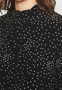 Zwarte blouse met witte stippen, voorzien van een hoge kraag en subtiele knooplijstdetails. Lichtgewicht stof met een gladde textuur.