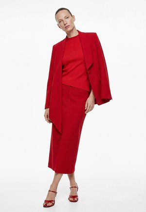 Femme portant un blazer rouge posé sur les épaules, un haut rouge assorti et une jupe midi, associée à des sandales rouges à lanières, posant devant un fond blanc.