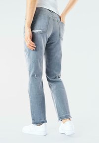 Jeans de mezclilla gris con un corte relajado, que presentan detalles desgastados, dos bolsillos traseros y dobladillos deshilachados. Combinados con zapatillas blancas.
