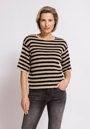 Woman with short blonde hair wearing a beige and black striped short-sleeve sweater and dark gray jeans standing against a white background.