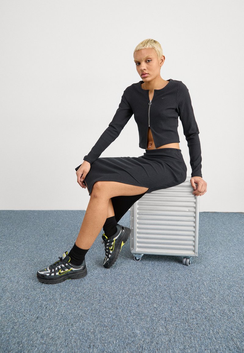 Chaqueta corta de punto acanalado negra con cremallera frontal, acompañada de una falda midi a juego con una abertura lateral. Completado con zapatillas deportivas negras.