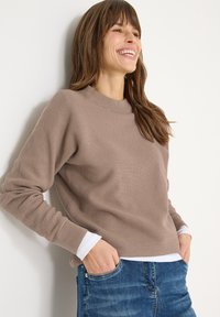 Brązowy prążkowany sweter z okrągłym dekoltem. Posiada długie rękawy i luźny krój, zestawiony z białą podkoszulką i niebieskimi dżinsami.