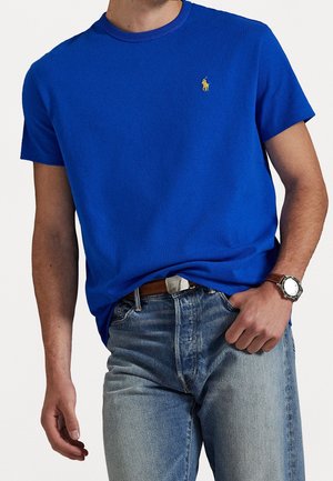 Mann trägt ein hellblaues kurzärmeliges T-Shirt mit einem kleinen gelben gestickten Logo, hellblaue Jeans, braunen Gürtel und Armbanduhr.