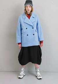 Manteau croisé en laine bleu clair avec boutons noirs, associé à une jupe plissée noire et des baskets métalliques argentées.