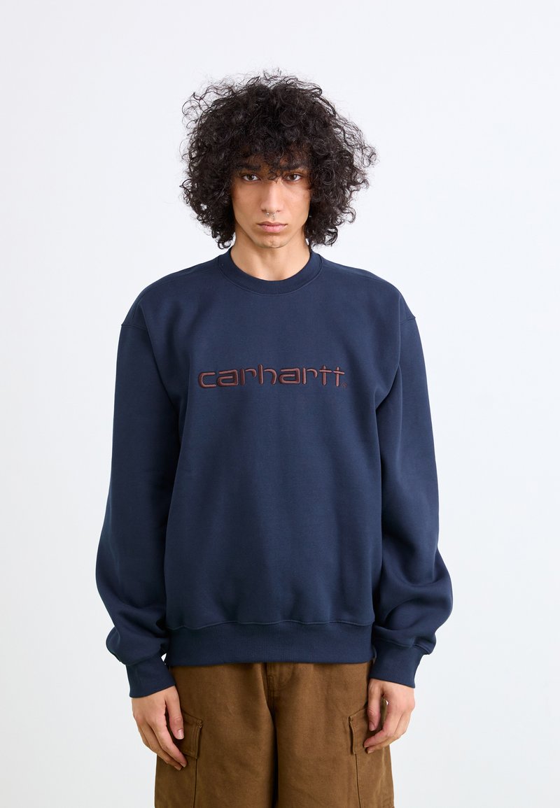 Navy blue sweatshirt with a crew neck, featuring a brown embroidered "carhartt" logo centered on the chest, made from textured fabric.