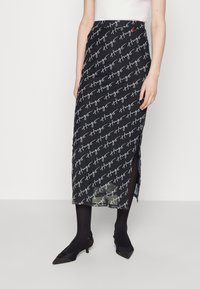 Falda midi negra con un patrón repetitivo de firma blanca, abertura lateral y un diseño ajustado, llevada sobre medias negras y tacones.