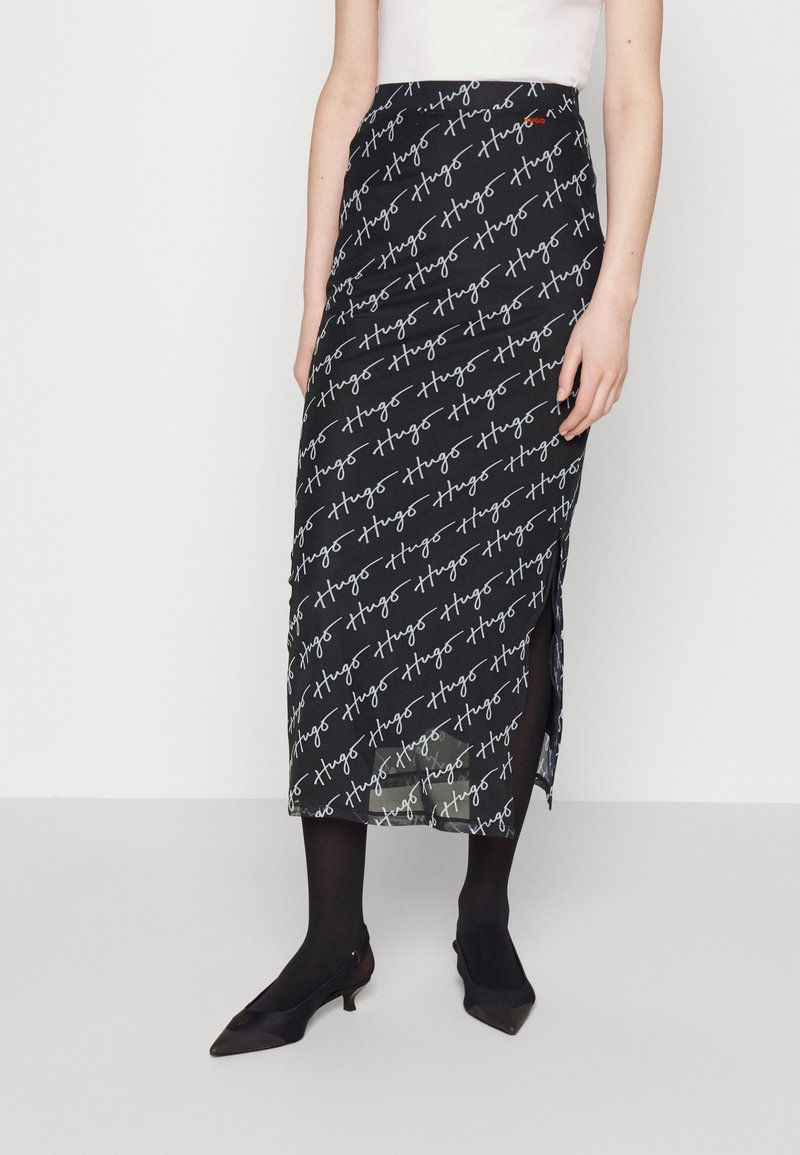 Falda midi negra con un patrón repetitivo de firma blanca, abertura lateral y un diseño ajustado, llevada sobre medias negras y tacones.