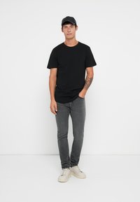 Camiseta negra de manga corta combinada con unos vaqueros gris ajustados y zapatillas blancas. El modelo lleva una gorra negra y tiene un tatuaje en el brazo.