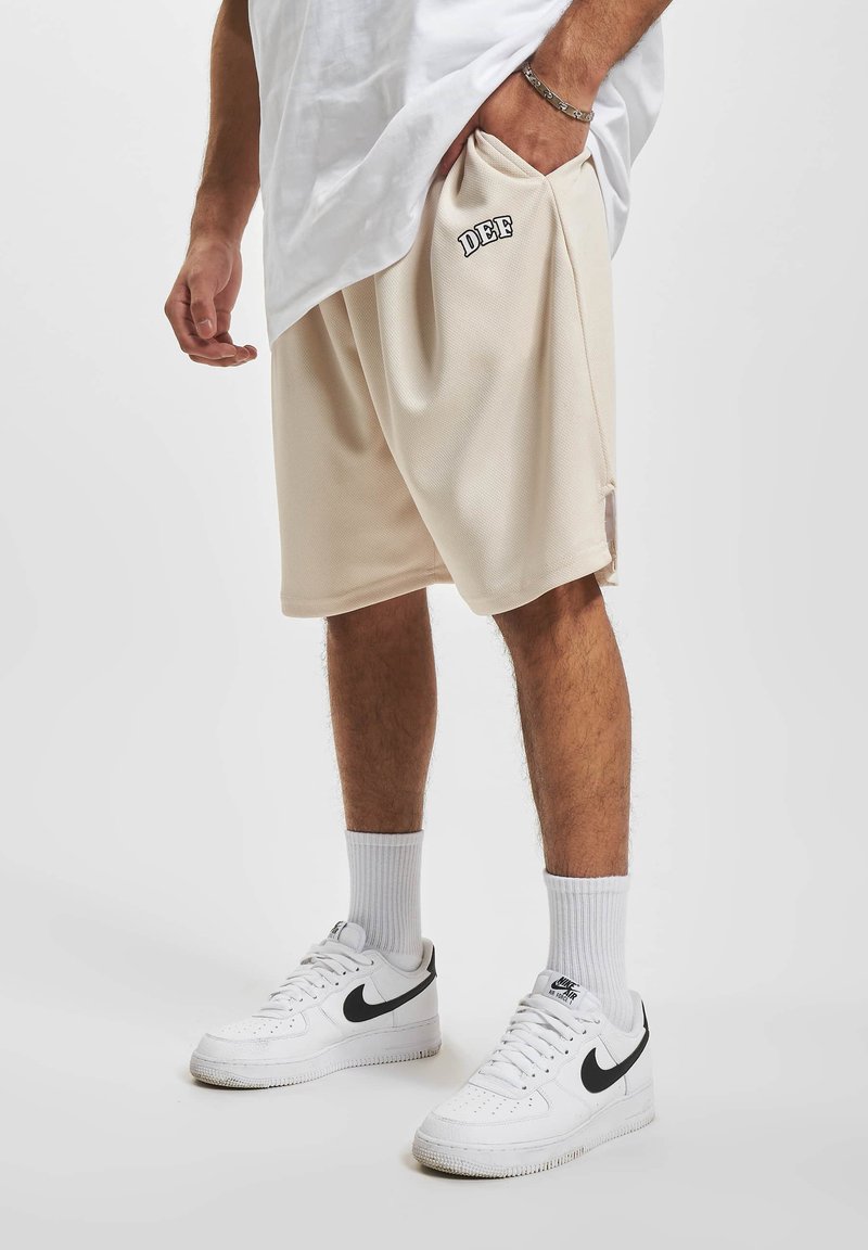 Shorts en maille beige à coupe loose, arborant un graphisme "DEF" sur le côté. Associés à des baskets blanches et des chaussettes blanches côtelées.