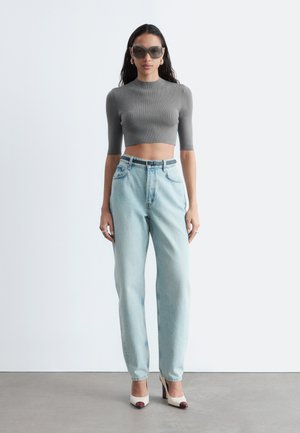 Donna che indossa un top grigio corto a costine, jeans azzurri a vita alta con cintura nera, tacchi bianchi con punte marroni e grandi occhiali da sole.