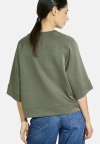 Sweatshirt vert surdimensionné avec un col rond, des manches courtes raglan et une coupe décontractée. Texture lisse avec des détails de coutures visibles.