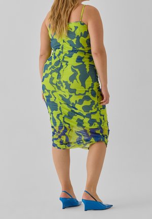 Femme portant une robe midi froncée à motif abstrait vert et bleu avec fines bretelles et chaussures slingback bleues à petit talon compensé.