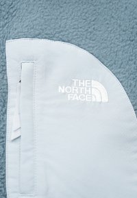 Κοντινό πλάνο από ανοιχτό μπλε φλις ύφασμα με μια ανοιχτό μπλε επιφάνεια που δείχνει το λογότυπο "THE NORTH FACE" κεντημένο με λευκή κλωστή.