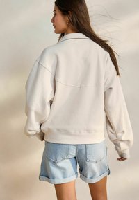 Felpa oversize color beige chiaro con spalle cadenti e polsini a coste, abbinata a pantaloni shorts in denim chiaro con orli arrotolati.