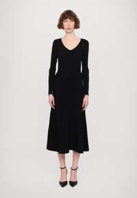 Abito lavorato a maglia nero con scollatura a V e maniche lunghe, corpetto aderente, gonna svasata e tacchi con cinturino alla caviglia. Tessuto liscio e colore solido.