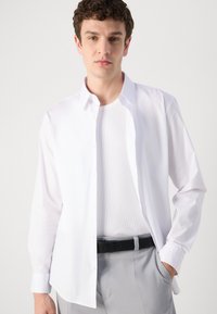 Weißes Langarm-Hemd mit Knopfleiste, das einen klassischen Kragen und eine glatte Textur aufweist, über ein weißes geripptes Tanktop getragen. Hellgraue Hose mit schwarzem Gürtel.