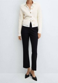 Kardigan w kolorze kremowym, fakturowany, zapinany na guziki, nałożony na białą koszulę, w połączeniu z dopasowanymi czarnymi spodniami i czarnymi balerinami z szpiczastym noskiem.