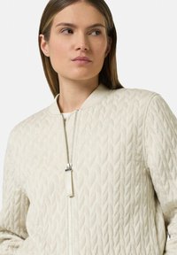Quilted light beige bomber jacket with a stand-up collar, silver zipper, and textured leaf pattern. Smooth fabric and fitted design.