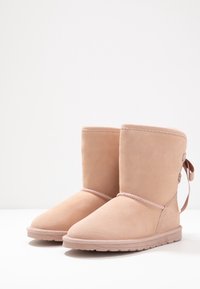 Botas de ante rosa claro hasta el tobillo con punta redondeada, costuras decorativas y un lazo en la parte posterior. Suelas de goma planas.